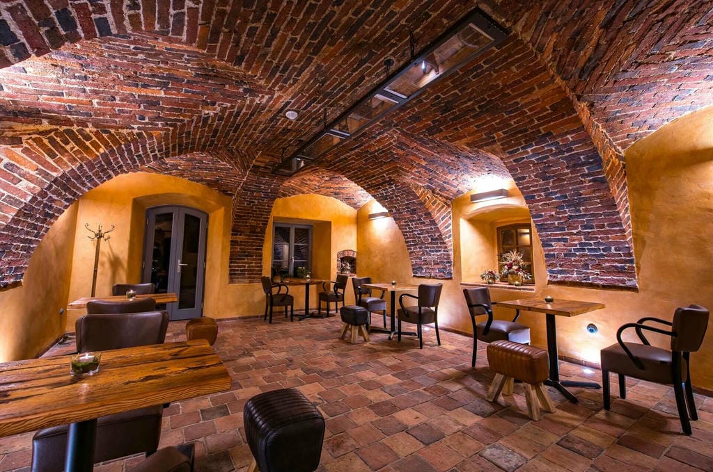 Interiér kavárny s dřevěnými stoly a koženými křesly v části s historickými klenbami.