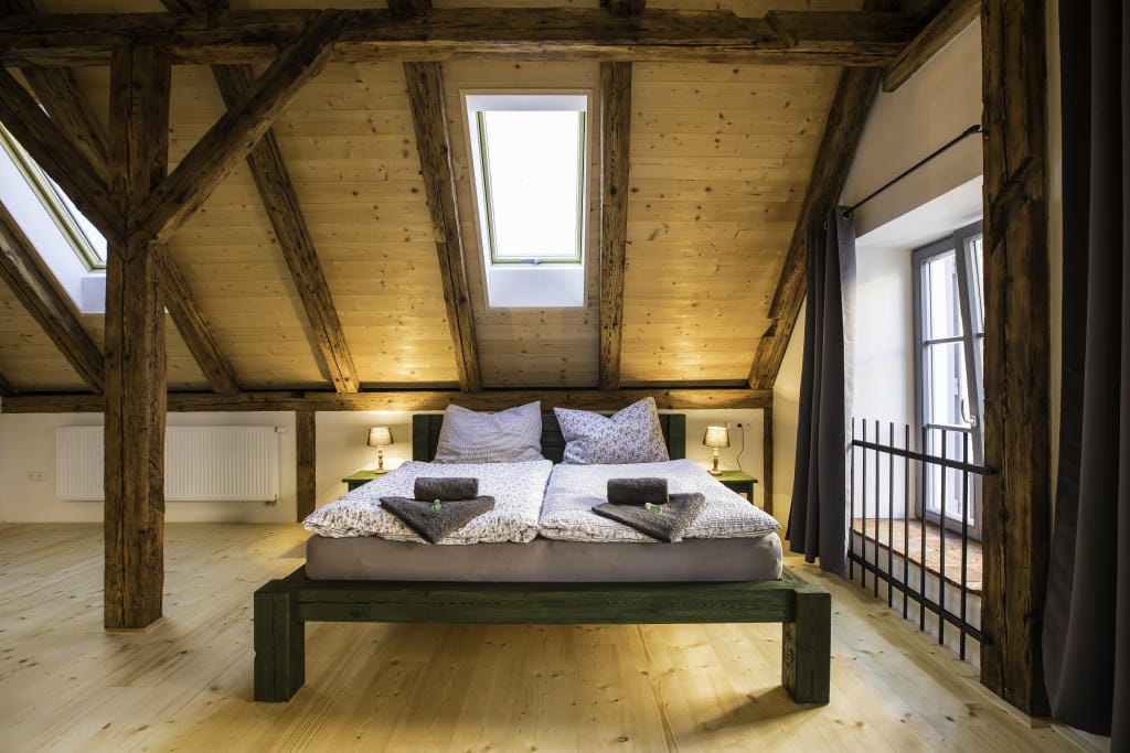 Manželská postel z masivu pod střešním oknem v ložnici s přiznanými dřevěnými trámy a světlou podlahou.